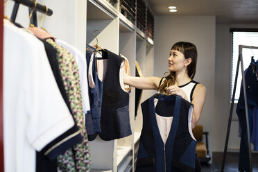 Japanese saleswoman standing in clothing store, hanging blue waistcoats on rail. - MINF09678