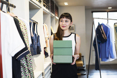 Eine japanische Verkäuferin steht in einem Bekleidungsgeschäft und hält einen Stapel grüner Kartons. - MINF09679
