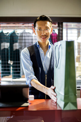 Japanischer Verkäufer mit Schnurrbart und Brille steht an der Kasse eines Bekleidungsgeschäfts, hält eine grüne Einkaufstasche und lächelt in die Kamera., lizenzfreies Stockfoto