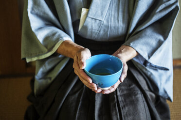 Nahaufnahme eines japanischen Mannes in traditionellem Kimono, der während einer Teezeremonie auf dem Boden kniet und eine blaue Teeschale hält. - MINF09682