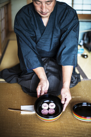 Japanischer Mann im traditionellen Kimono kniet auf dem Boden und hält eine Schale mit Wagashi, Süßigkeiten, die traditionell während einer japanischen Teezeremonie serviert werden., lizenzfreies Stockfoto