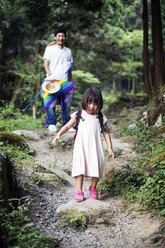 Japanese girl wearing pale pink sun dress and carrying backpack standing in a forest, man in the background. - MINF09701