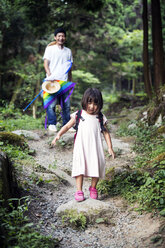 Japanisches Mädchen in blassrosa Sonnenkleid und mit Rucksack in einem Wald stehend, Mann im Hintergrund. - MINF09701