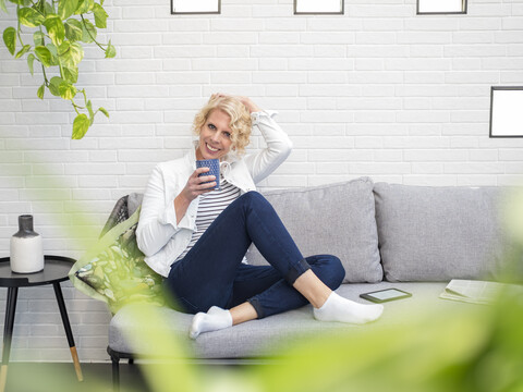Porträt einer lächelnden reifen Frau, die zu Hause auf der Couch sitzt und einen Kaffeebecher hält, lizenzfreies Stockfoto