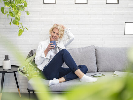 Porträt einer lächelnden reifen Frau, die zu Hause auf der Couch sitzt und einen Kaffeebecher hält - LAF02170