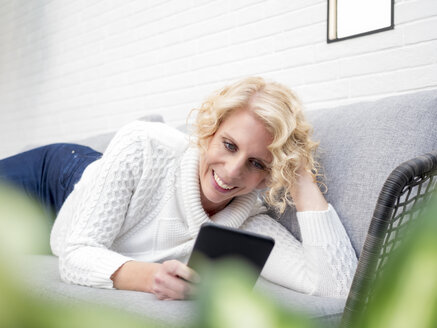 Lächelnde reife Frau liegt auf der Couch zu Hause und liest ein Buch - LAF02172