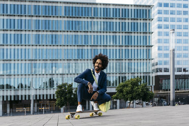 Spain, Barcelona, young businessman crouching on skateboard in the city - JRFF02040