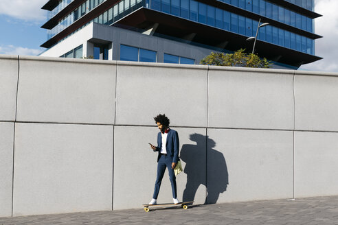 Spain, Barcelona, young businessman riding skateboard and using cell phone in the city - JRFF02048