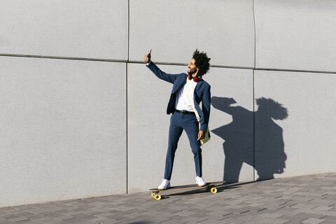 Junger Geschäftsmann fährt Skateboard entlang einer Wand und macht ein Selfie, lizenzfreies Stockfoto