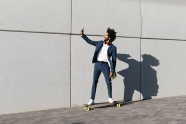 Junger Geschäftsmann fährt Skateboard entlang einer Wand und macht ein Selfie - JRFF02049