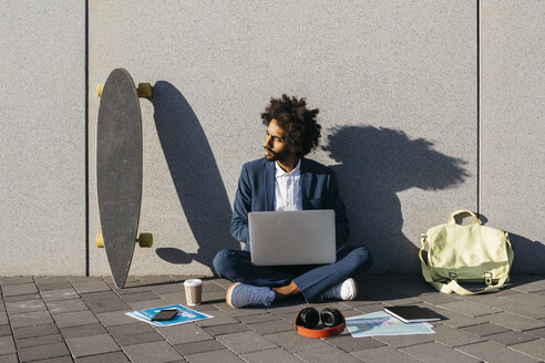 Junger Geschäftsmann sitzt im Freien an einer Wand und arbeitet an einem Laptop - JRFF02060