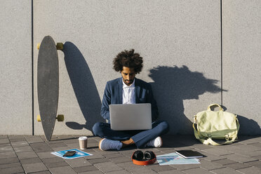 Young businessman sitting outdoors at a wall working on laptop - JRFF02061