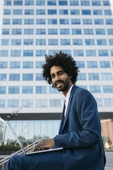 Spain, Barcelona, smiling young businessman sitting outdoors in the city working on laptop - JRFF02066