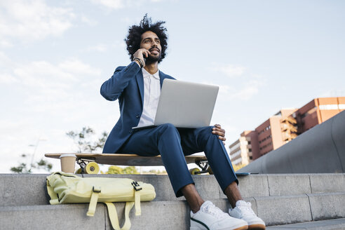 Spain, Barcelona, young businessman sitting outdoors in the city using cell phone and laptop - JRFF02067