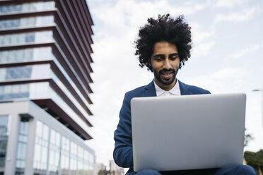 Spain, Barcelona, smiling young businessman sitting outdoors in the city working on laptop - JRFF02070