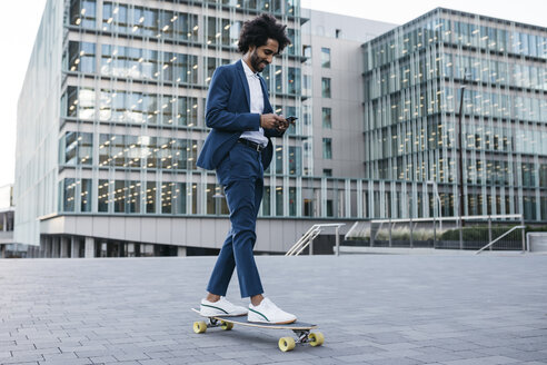 Spain, Barcelona, young businessman riding skateboard and using cell phone in the city - JRFF02079