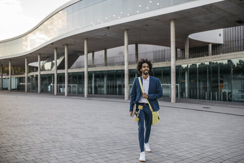 Spain, Barcelona, young businessman leaving office building holding skateboard in the city - JRFF02080