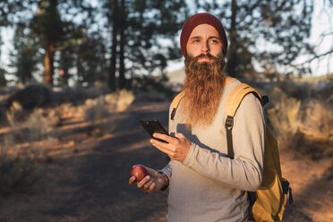 USA, Nordkalifornien, bärtiger Mann mit Handy und Apfel in einem Wald in der Nähe des Lassen Volcanic National Park - KKAF02980