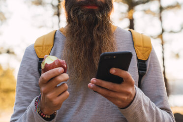 Nahaufnahme eines bärtigen Mannes, der ein Handy benutzt und einen Apfel bei einer Wanderung im Wald isst - KKAF02984