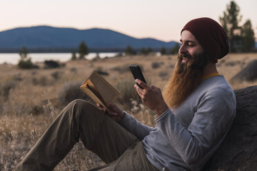 USA, North California, bearded young man with book and cell phone during a hiking trip near Lassen Volcanic National Park - KKAF02996