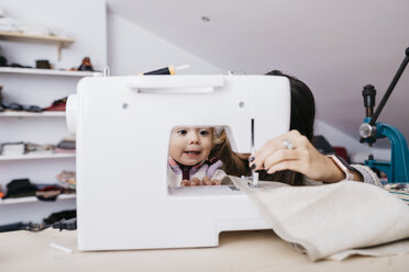 Little daughter watching her mother using sewing machine at home - JRFF02085
