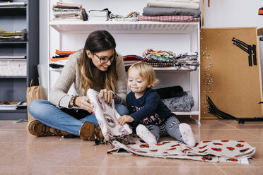 Mutter und kleine Tochter sitzen zu Hause auf dem Boden und prüfen Stoffe - JRFF02094