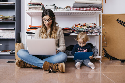 Mutter und kleine Tochter sitzen zu Hause auf dem Boden und benutzen Laptop und Tablet, lizenzfreies Stockfoto