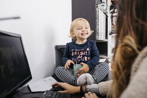 Mutter arbeitet zu Hause mit kleiner Tochter auf dem Schreibtisch sitzend, lizenzfreies Stockfoto