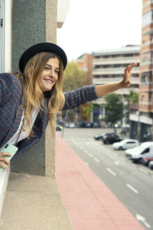 Smiling young woman leaning out of window waving - ERRF00097