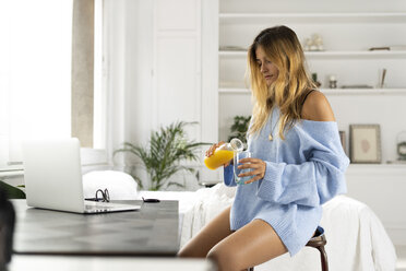 Junge Frau sitzt im Schlafzimmer zu Hause und gießt Orangensaft in ein Glas - ERRF00106
