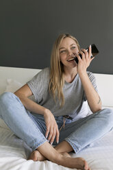 Glückliche junge Frau, die auf dem Bett sitzt und ein Mobiltelefon benutzt - VABF01714