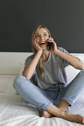 Glückliche junge Frau sitzt auf dem Bett und spricht mit ihrem Handy - VABF01715