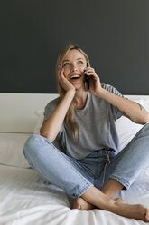 Happy young woman sitting on bed talking on cell phone - VABF01715