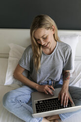 Glückliche junge Frau, die auf dem Bett sitzt und einen Laptop benutzt - VABF01716