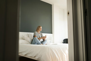 Junge Frau sitzt auf dem Bett und benutzt ein Mobiltelefon - VABF01717