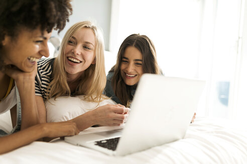 Drei glückliche Freundinnen liegen auf dem Bett und teilen sich einen Laptop - VABF01723