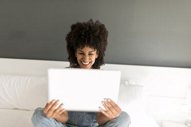 Happy woman sitting on bed looking at tablet - VABF01755