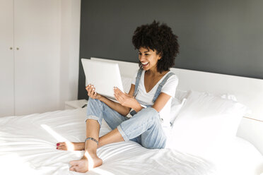 Happy woman sitting on bed looking at tablet - VABF01757