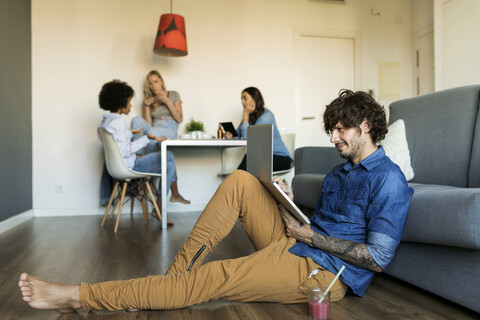 Smiling man sitting on floor using laptop with friends in background stock photo
