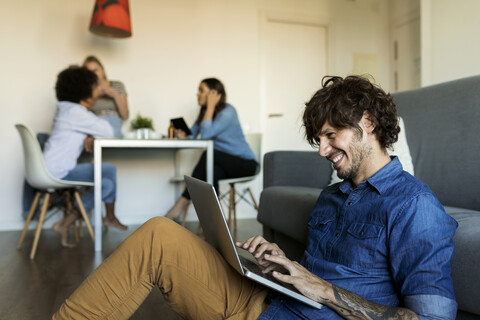 Lächelnder Mann sitzt auf dem Boden und benutzt einen Laptop mit Freunden im Hintergrund, lizenzfreies Stockfoto