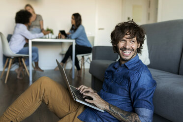 Lachender Mann sitzt auf dem Boden und benutzt einen Laptop mit Freunden im Hintergrund - VABF01766