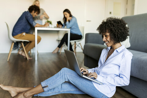 Smiling woman sitting on floor using laptop with friends in background stock photo