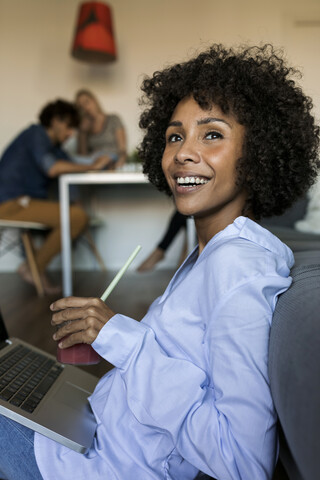 Glückliche Frau mit einem alkoholfreien Getränk auf dem Boden sitzend und mit einem Laptop und Freunden im Hintergrund, lizenzfreies Stockfoto