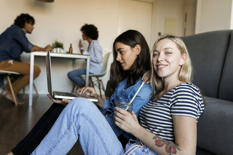 Zwei lächelnde junge Frauen sitzen auf dem Boden und teilen sich einen Laptop mit Freunden im Hintergrund, lizenzfreies Stockfoto