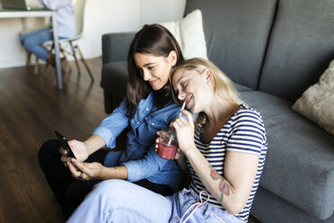 Zwei lächelnde junge Frauen sitzen mit einem Softdrink auf dem Boden und teilen sich ein Mobiltelefon - VABF01777