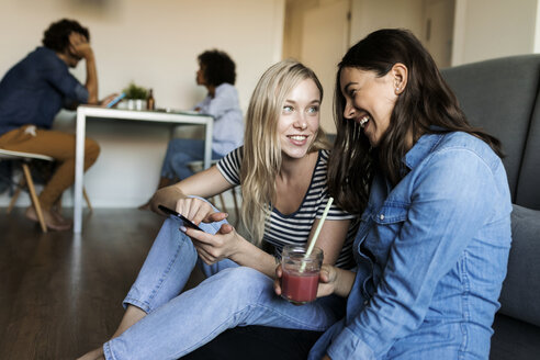 Zwei fröhliche junge Frauen sitzen auf dem Boden mit Handy und Getränk und Freunde im Hintergrund - VABF01779
