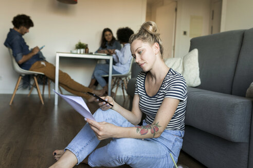 Junge Frau sitzt auf dem Boden mit Handy und Papieren und Freunden im Hintergrund - VABF01780