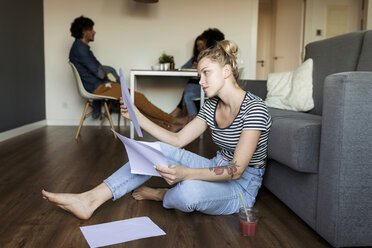 Junge Frau sitzt auf dem Boden mit Papieren und Freunden im Hintergrund - VABF01783