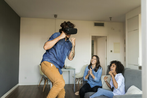 Zwei Frauen ermutigen Mann beim Spielen mit VR-Brille zu Hause, lizenzfreies Stockfoto