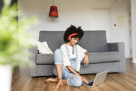 Lächelnde Frau sitzt zu Hause mit Kopfhörern und Laptop, lizenzfreies Stockfoto